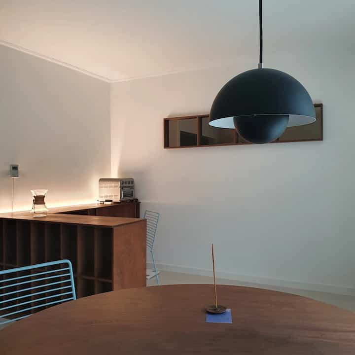 白い壁と木目調のテーブルがあるミニマルでナチュラルモダンなダイニング、中央に大きな黒いペンダントライトが配置された印象的な空間。