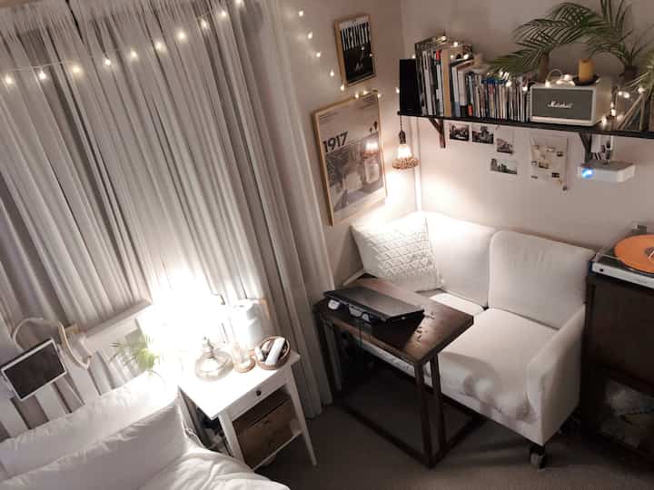 A cozy white and natural tone studio bedroom featuring a white sofa, wall shelf with books, and a mounted projector for home cinema ambiance