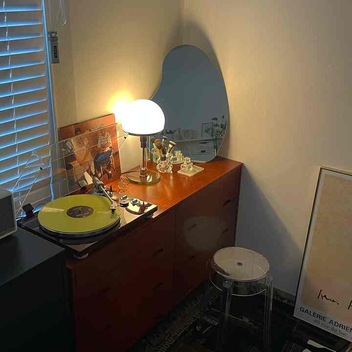 Cozy living room corner with warm beige and brown tones, wooden dresser topped with a lamp and transparent stool, unique mirror and poster displayed