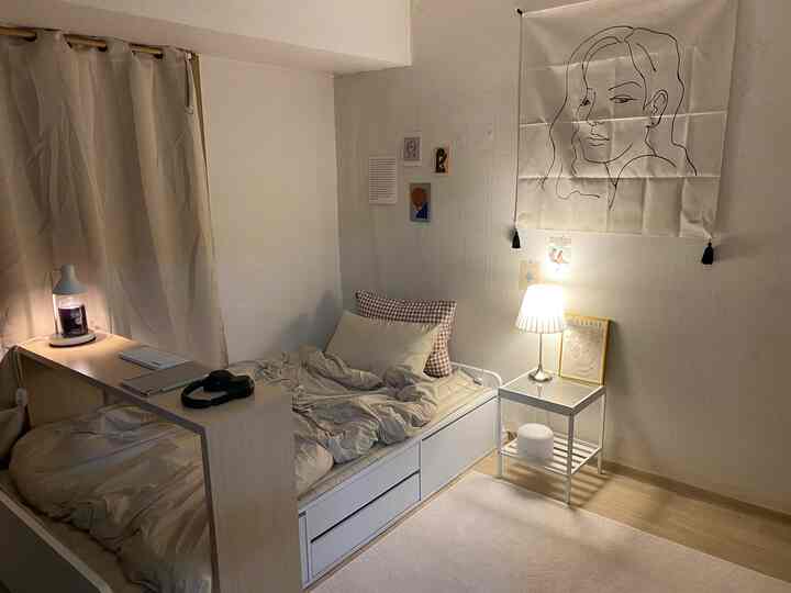 A small bedroom in white and beige tones featuring a single bed and minimal nightstand with warm lighting creating a cozy atmosphere