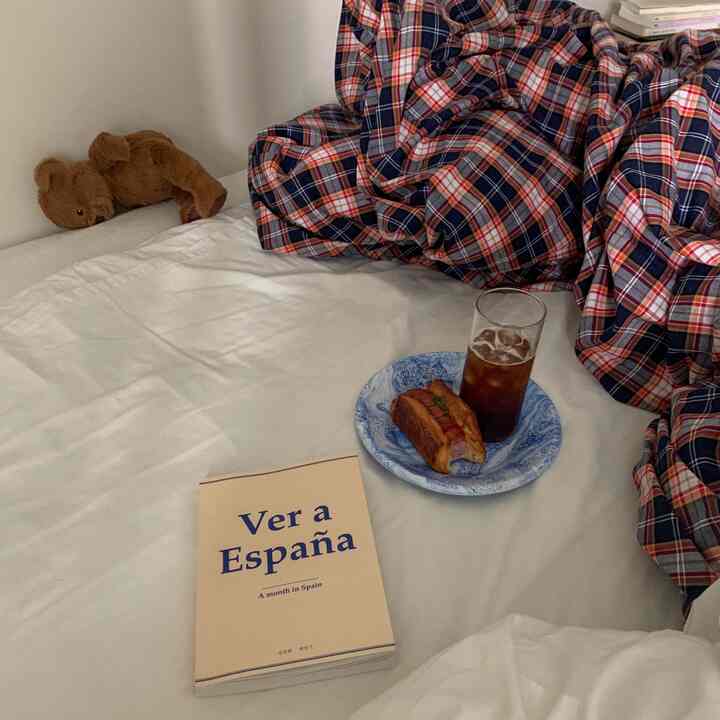 白い寝具の寝室にネイビーのチェック掛け布団、ぬいぐるみのクマと飲み物・パンが載った皿が配置され、居心地の良いおうち時間の雰囲気を醸し出す