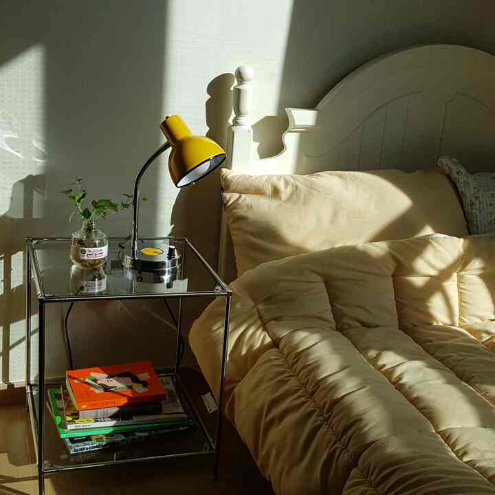 Bright bedroom with white wooden bed featuring beige bedding and a yellow lamp on a glass side table, creating a warm natural ambiance