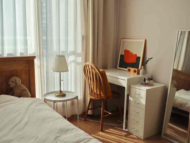 A warm-toned single household bedroom home office featuring white curtains, wood-toned desk and bed with cozy atmosphere