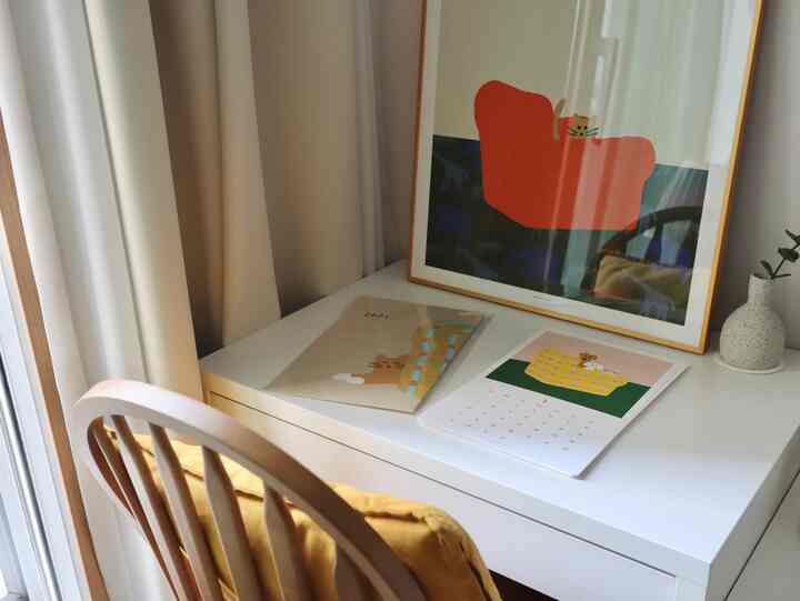 Bright white toned natural style home office space featuring white desk with calendars, a framed poster, and a yellow cushioned chair creating a cozy atmosphere