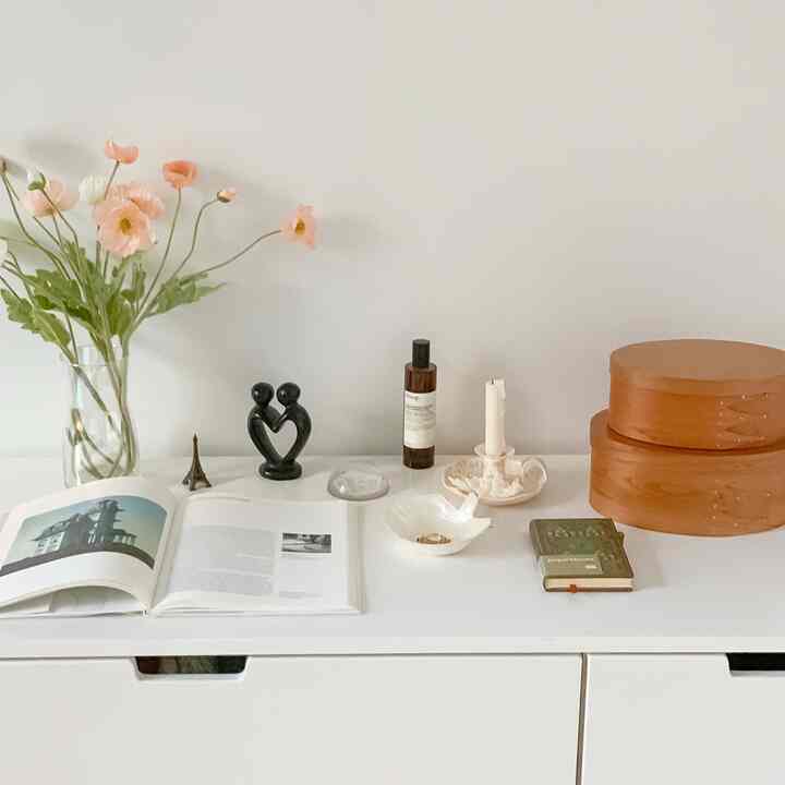 A white-toned cabinet topped with wood-tone objets, a vase of flowers, and books in a simple, minimalist interior space
