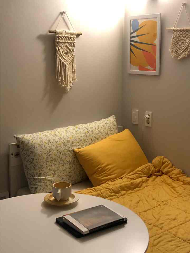 Cozy small single bedroom in warm yellow and beige tones, featuring a Nordic style bed with cushions and mattress, macrame wall decor and abstract framed art