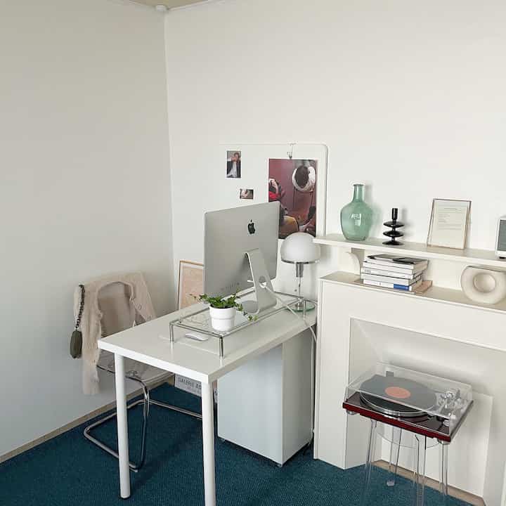 White-walled studio home office featuring a white desk, transparent stool, and a decorative fireplace creating a clean modern vibe