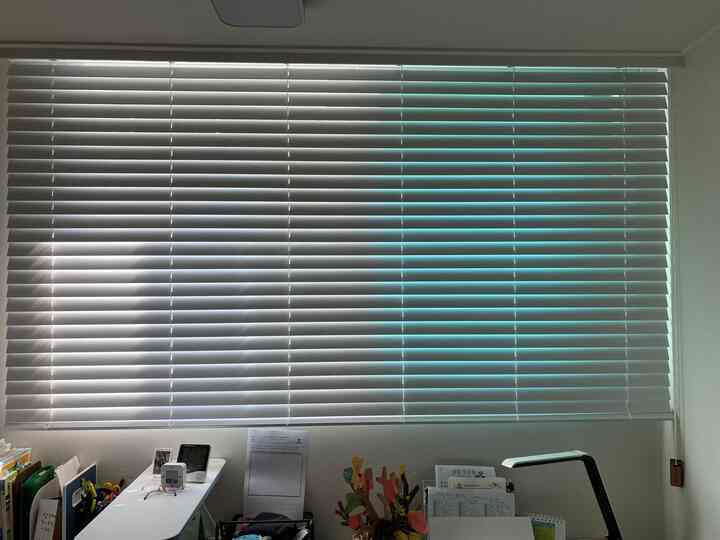 白を基調とした子供部屋、白いウッド調ブラインドが窓に設置され、下部には子供用机と文具が整然と配置されたシンプルな空間