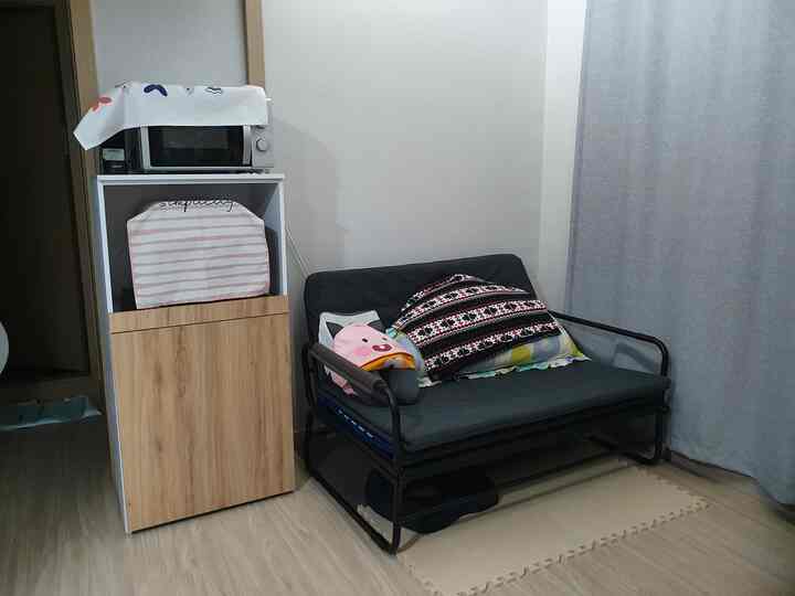Small room with gray curtains and natural wood-tone flooring, featuring a black sofa bed with assorted cushions creating a cozy atmosphere