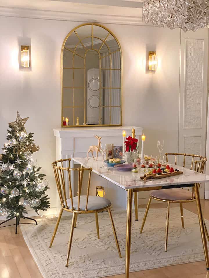A white and gold-toned living room featuring a Christmas tree, dining table with chairs, and wall sconces creating a warm home party atmosphere