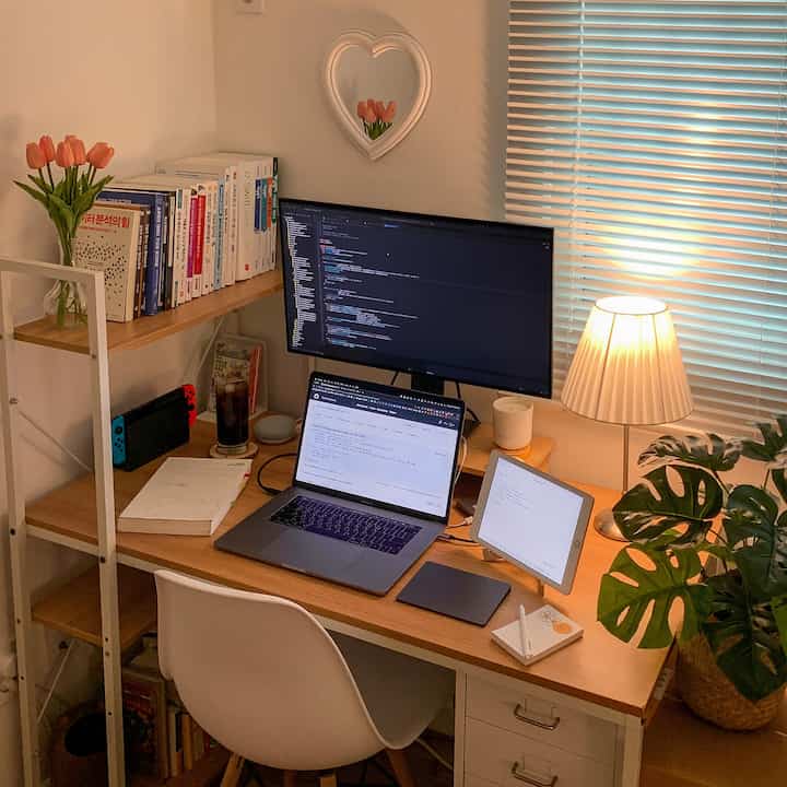 Bright white-walled home office featuring a wood-tone desk with monitor, laptop, and warm lighting creating a cozy workspace.