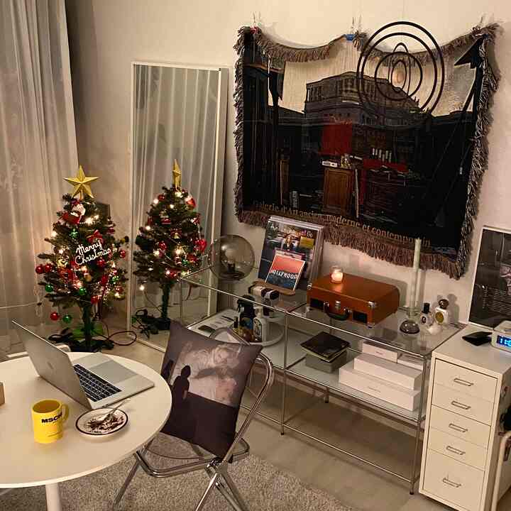 暖かい照明のモダンで白い小部屋、クリスマスツリーと透明なガラスのデスク、ノートパソコンとコーヒーマグが配置された居心地の良い在宅勤務空間