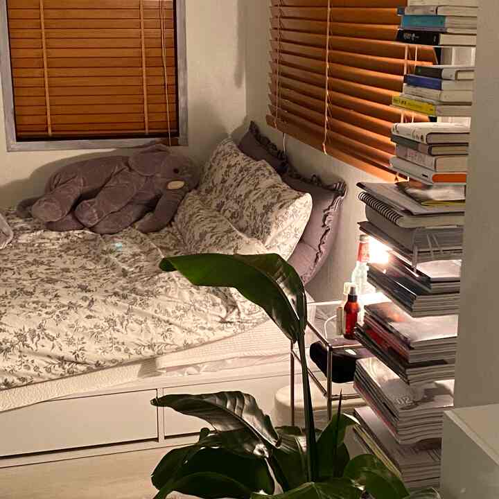A bedroom with white walls and wood tone blinds, featuring floral bedding and a stacked bookshelf creating a warm atmosphere