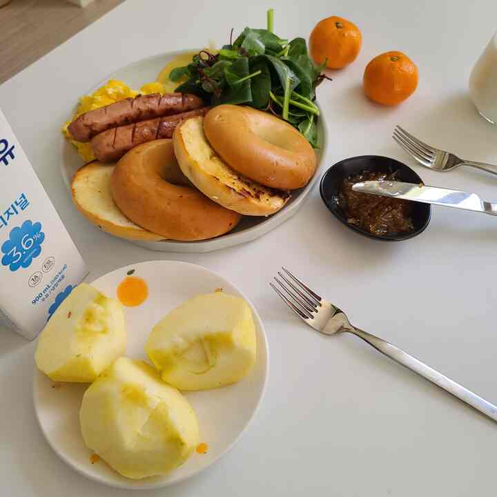 Simple brunch setting on a white dining table featuring bagels, sausages, salad, sliced apples, and a stone sauce bowl