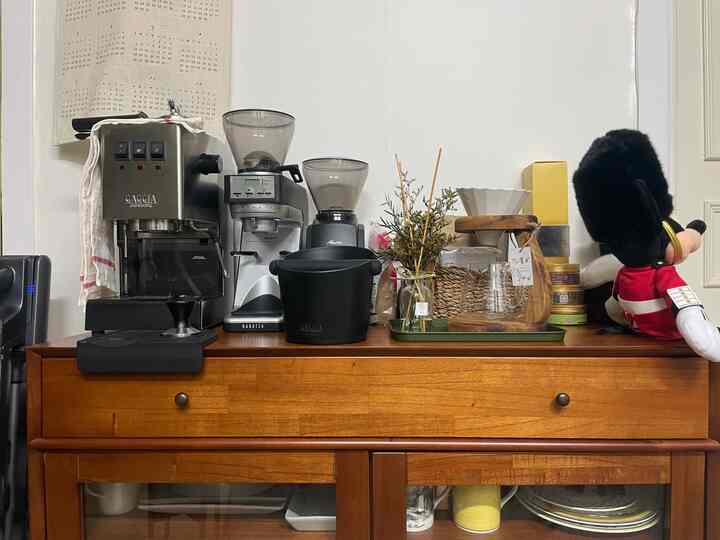 Warm brown-toned kitchen corner featuring home cafe setup with coffee machines, grinders, diffuser, and glassware on wooden cabinet