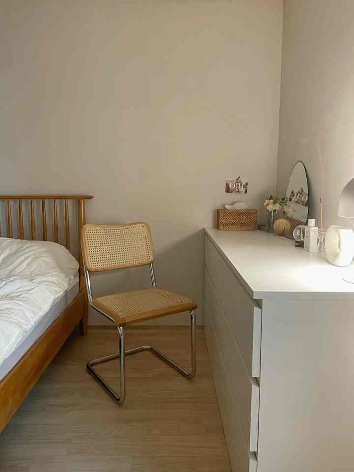 Beige-toned bedroom featuring a wooden bed, rattan chair, white dresser with round mirror, creating a simple and natural atmosphere