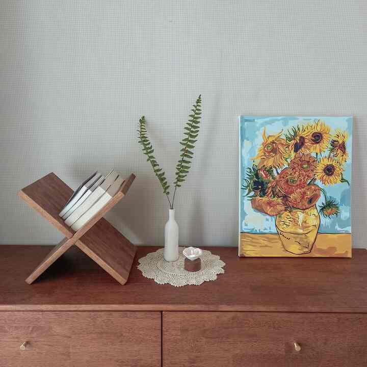 A cozy bedroom corner in warm brown and ivory tones featuring a wooden chest of drawers, bookshelf, ceramic vase, and sunflower painting