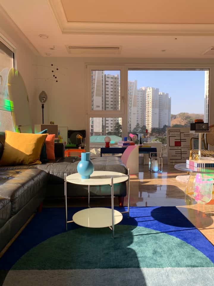 ナチュラルカラーとモダンな雰囲気の陽光あふれるリビング、黒いレザーソファと白いサイドテーブル、カラフルなラグが配置された印象的な空間