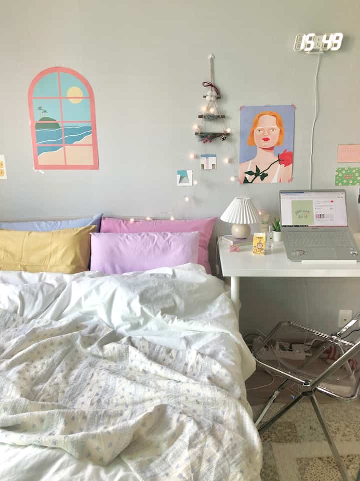Pastel-toned bedroom featuring a bed with pillows and throw, alongside a white side table with a laptop creating a cozy atmosphere