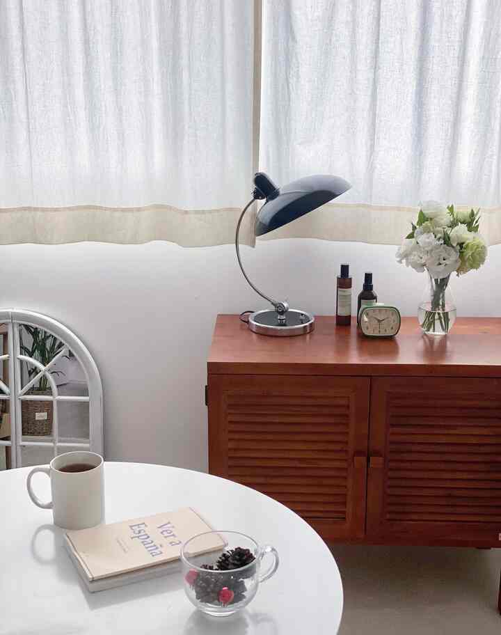 White walls and simple curtains background a living room corner with brown wooden console, table lamp, and vase of white flowers, evoking a cozy cafe style