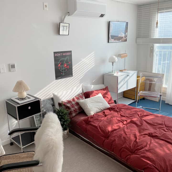 温かみのある赤い寝具が置かれたワンルーム寝室、モダンな家具と透明な椅子が調和する印象的な空間