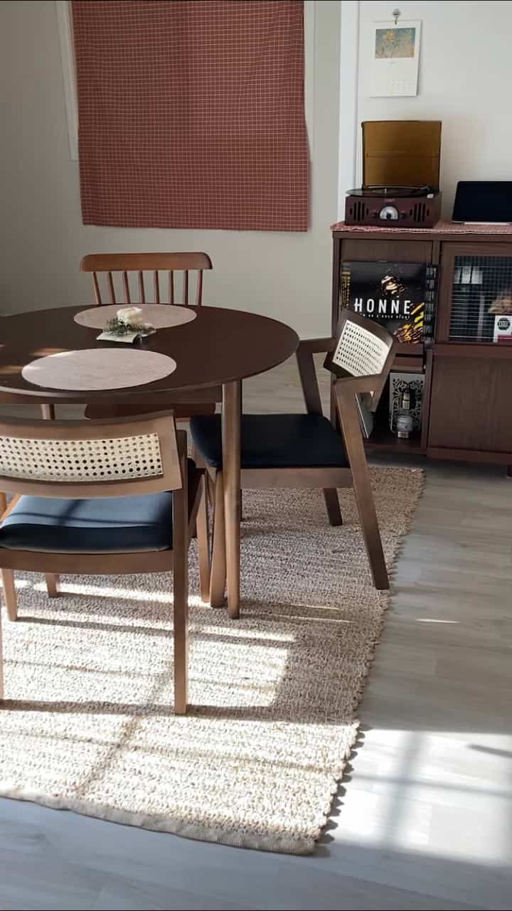 暖かみのあるブラウンの木目調のダイニングルーム、円形のダイニングテーブルとファブリックカーテン、明るい草ラグがカフェの雰囲気を演出する印象的な空間