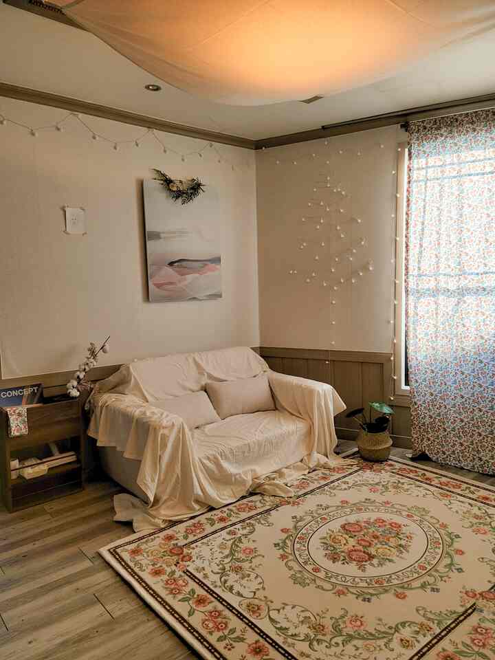 ベージュとブラウンのナチュラルトーンのリビング、白いカバーのソファと花柄のラグ、ラタンの鉢カバーが配置された雰囲気のある空間