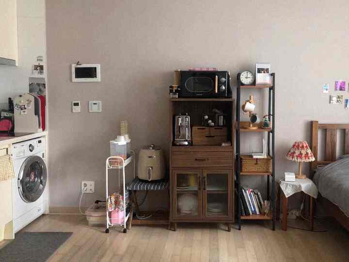Natural tone studio apartment featuring kitchen and bedroom with nightstand and coffee machines, cozy home cafe atmosphere in a compact room