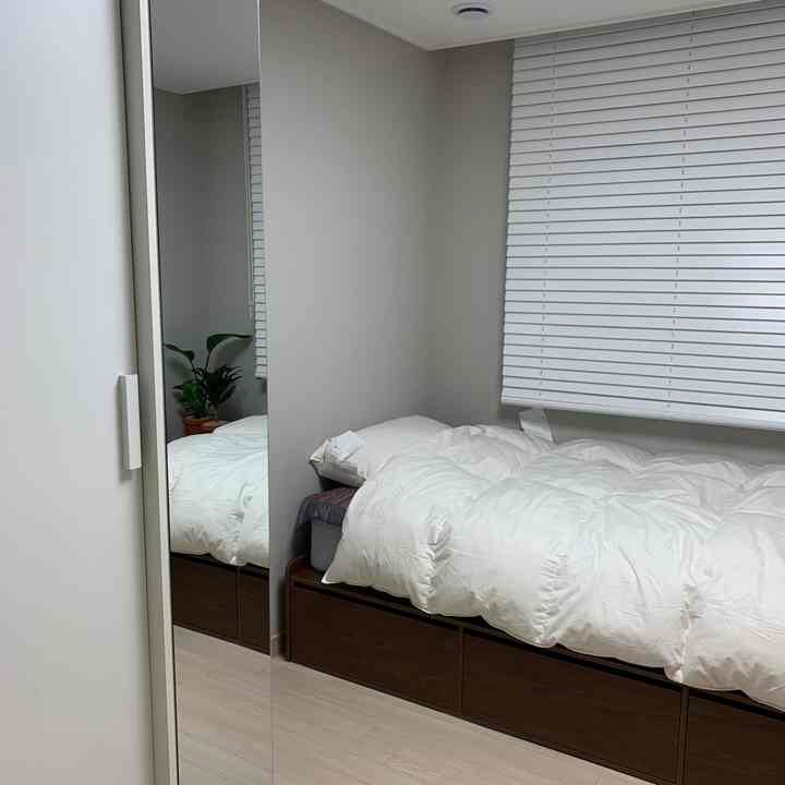 Clean modern bedroom with white walls and blinds, featuring wood-tone bed frame with storage in a simple setting