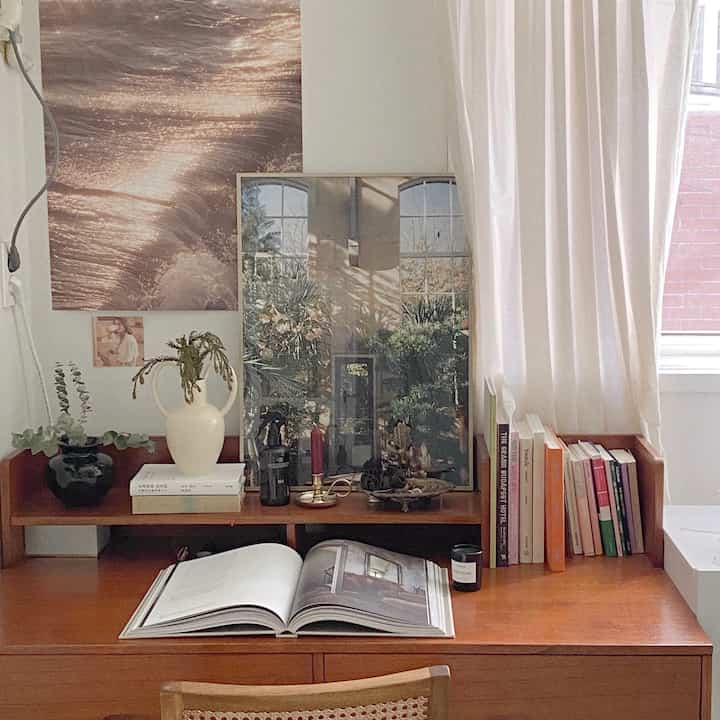 Natural wood-toned studio room workspace featuring a wooden desk, white curtains, and travel mood posters creating a cozy atmosphere