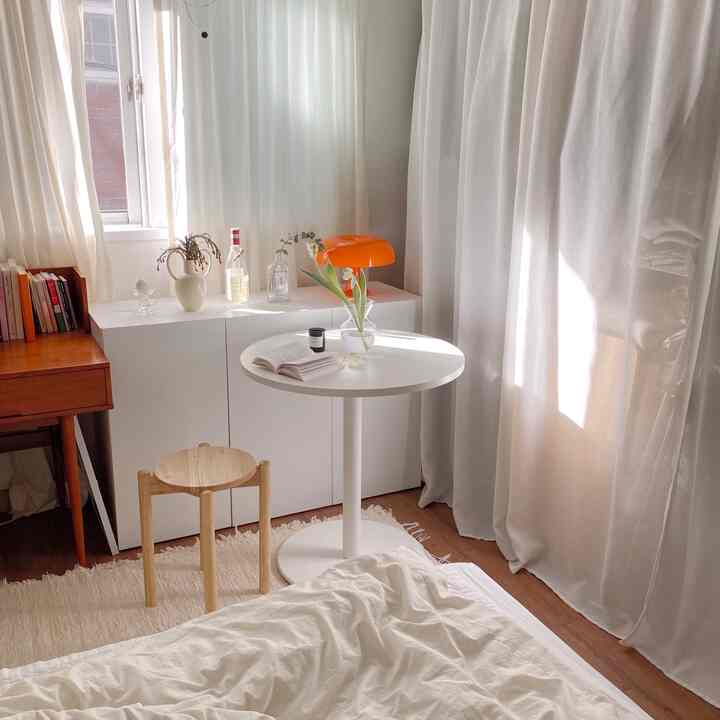 Natural and white toned studio apartment featuring round table, stool, bed, cabinet, and curtains creating a cozy atmosphere