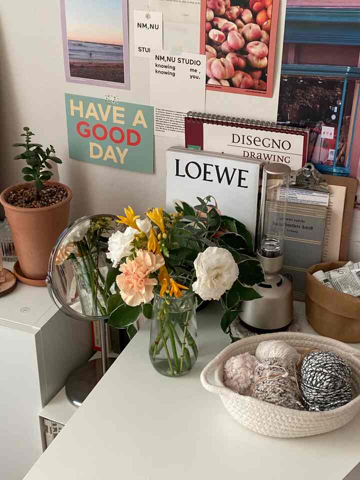 White and natural toned desk area featuring plants, flowers, and postcards, creating a simple and natural home office ambiance