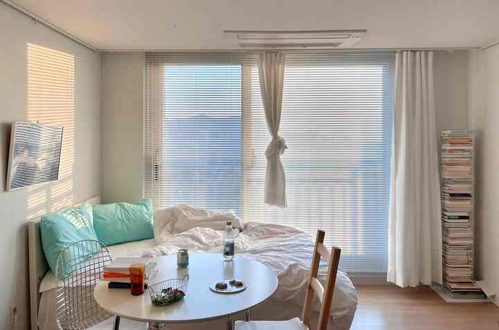 Bright white and natural wood-tone single household bedroom featuring round table, blinds, and stacked books for a clean and cozy ambiance