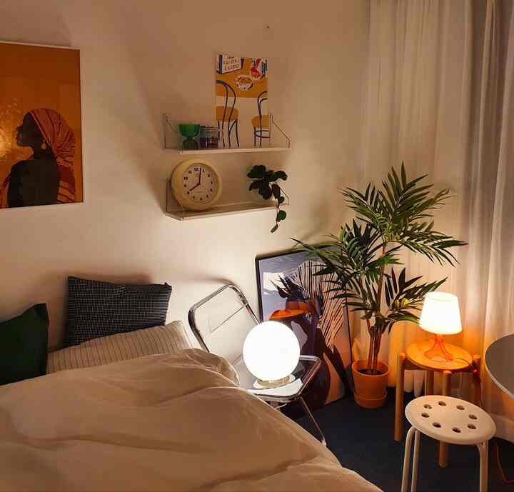 Beige and white toned bedroom featuring stools, transparent chair, multiple lights, and a large plant with cozy ambiance