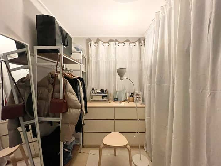 Small walk-in closet in white and natural tones featuring white curtains, dresser, full-length mirror, and stool with a neat atmosphere
