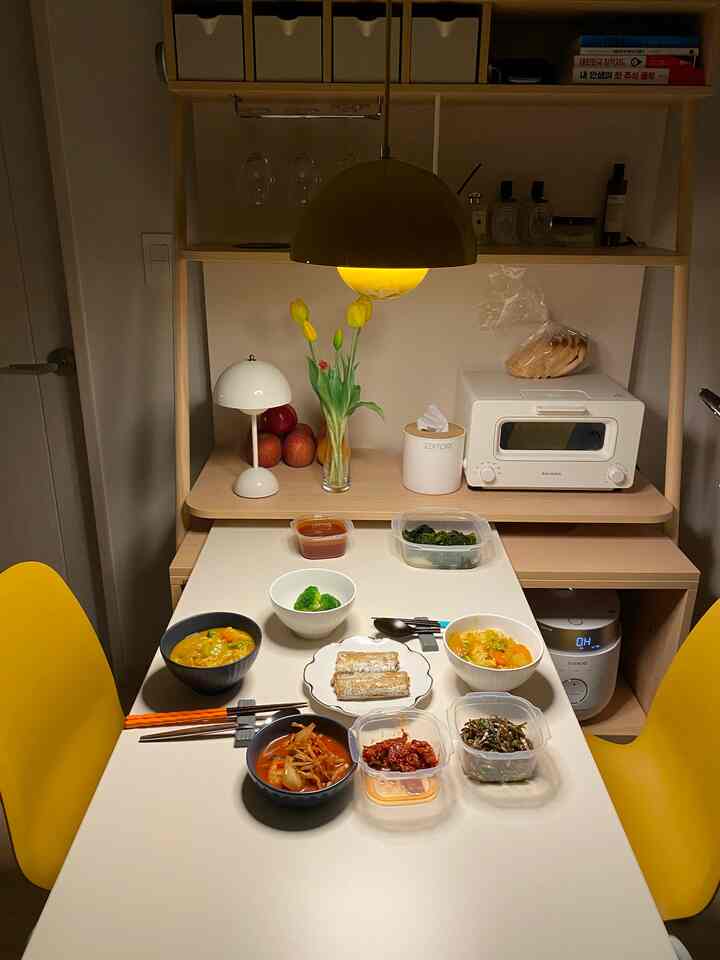 イエローのペンダントライトと椅子がある、ライトウッドの棚と収納が配置されたキッチンの落ち着いた空間