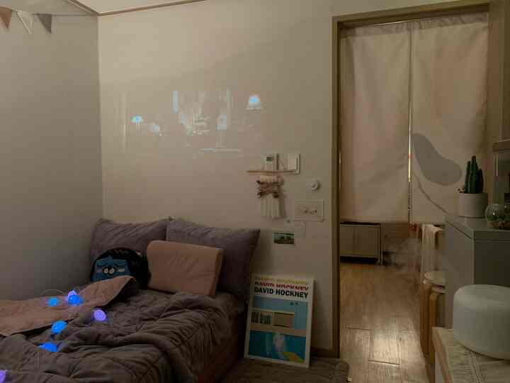 パープルとナチュラルブラウンのトーンのワンルーム寝室、ソファベッドと遮光カーテン、壁に投影されたホームシネマで落ち着いた雰囲気のインテリア