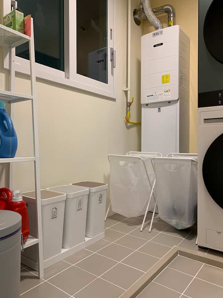 ベージュの落ち着いた色調の狭い多用途室、洗濯機と乾燥機、分別ゴミ箱、棚が配置された清潔な洗濯空間