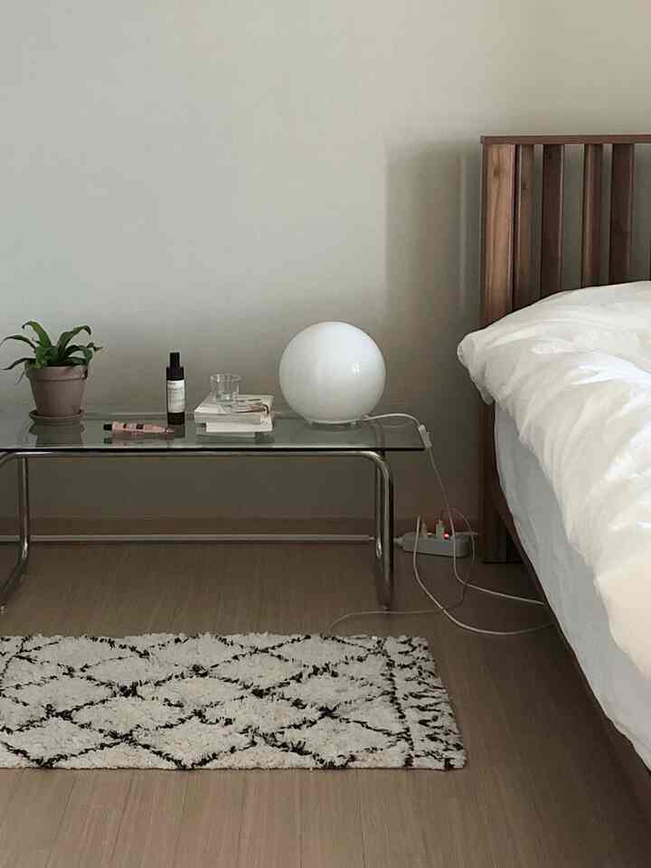 A bedroom in ivory and wood tones featuring a wooden bed frame, white bedding, a glass side table, and a patterned rug in a simple setting