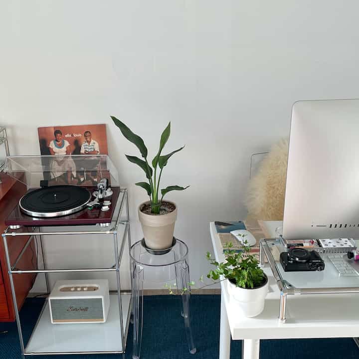 Minimal home office with white walls and blue carpet, featuring a transparent stool, vintage turntable, and green plants