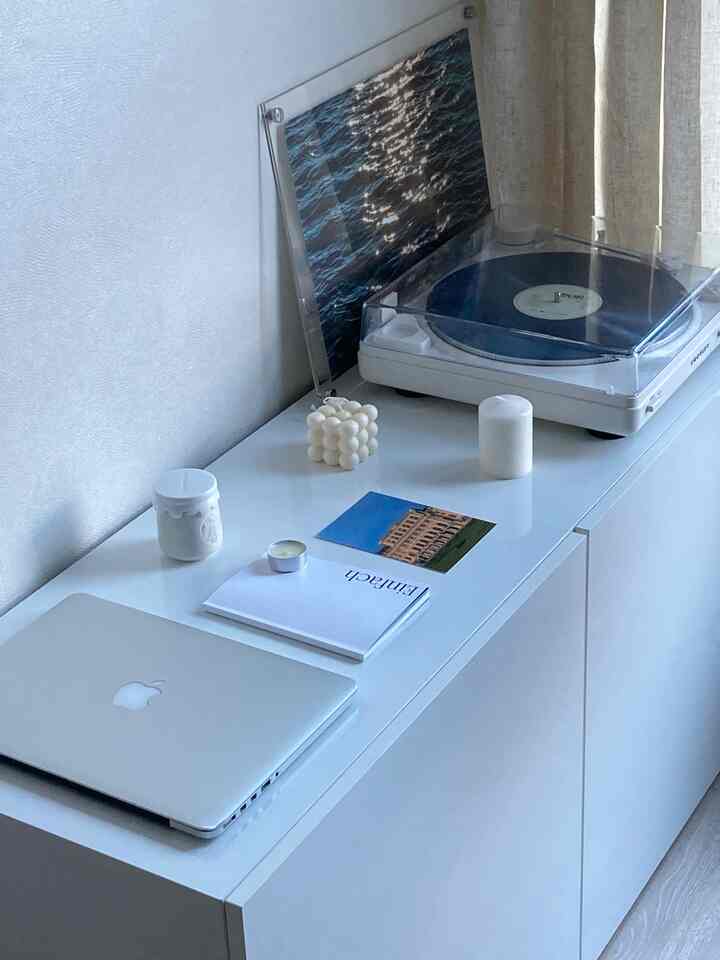 白を基調としたリビング空間、白いサイドボードの上にワイヤレスBluetoothターンテーブルとキャンドル、ノート、ノートパソコンが配置されたミニマルなインテリア
