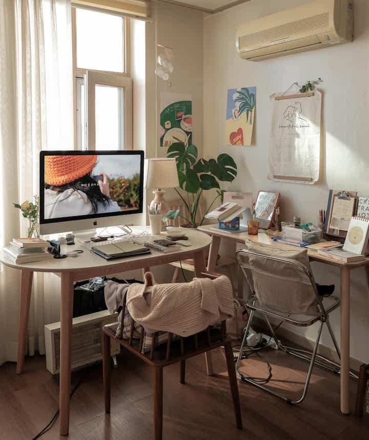 White and wood tone studio apartment workspace featuring two desks, curtains, and plants creating a simple and warm atmosphere