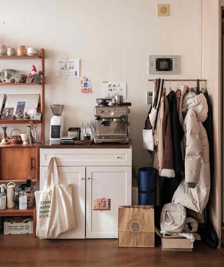 Studio apartment with white walls and brown wood flooring, featuring a home cafe setup with coffee equipment and a clothing rack in a cozy atmosphere