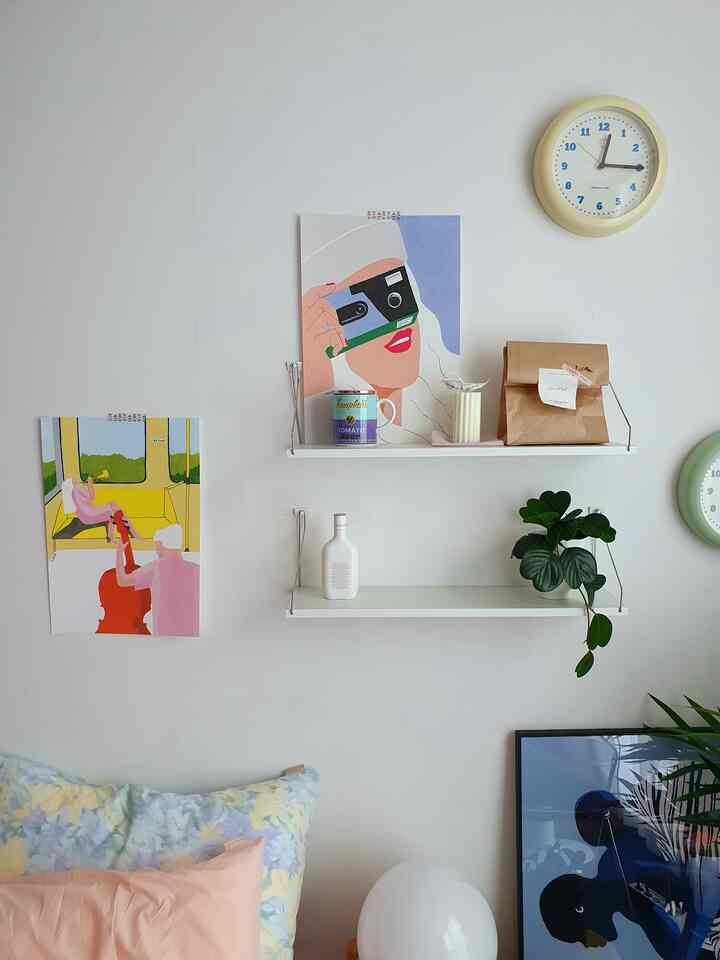 Living room wall with white backdrop and pastel pillows, decorated with posters, clocks, and wall shelves creating cozy ambiance
