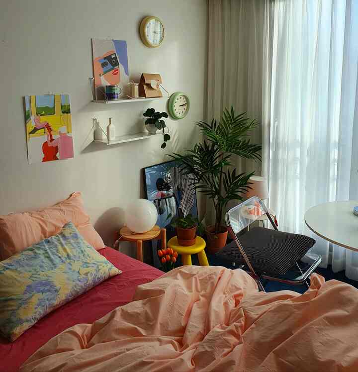 Cozy single bedroom with light pink bedding, wood-toned round furniture, and indoor plants bathed in soft natural light