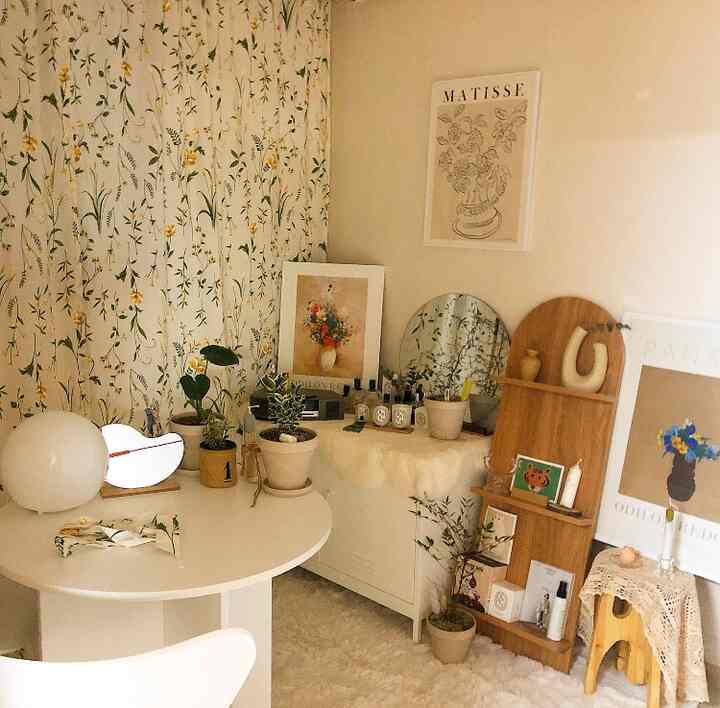 Beige and white toned small living room featuring a round table, floral curtains, various plants, and picture frames with a natural atmosphere