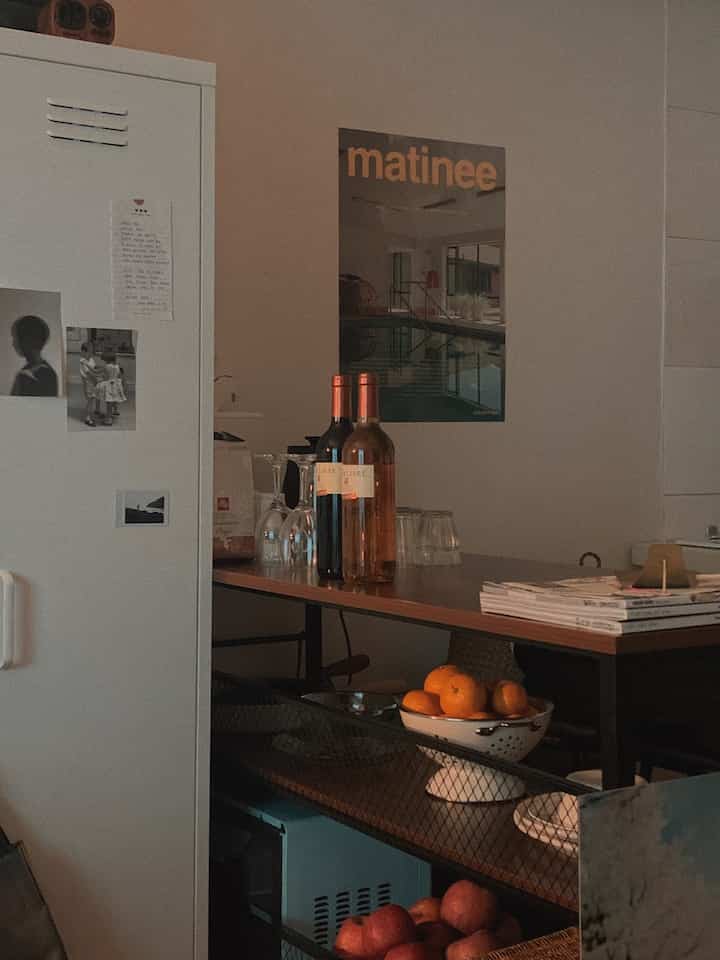 暖かいブラウンとホワイトのトーンのワンルームキッチン、中央にアイランドテーブルとワイングラス、ポスターが配された雰囲気のあるインテリア