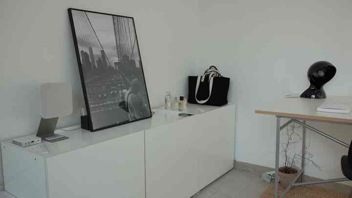 ホワイトトーンのシンプルな6畳のワンルーム、白い収納キャビネットとデスク、黒のテーブルランプと額縁が配置された雰囲気あるインテリア