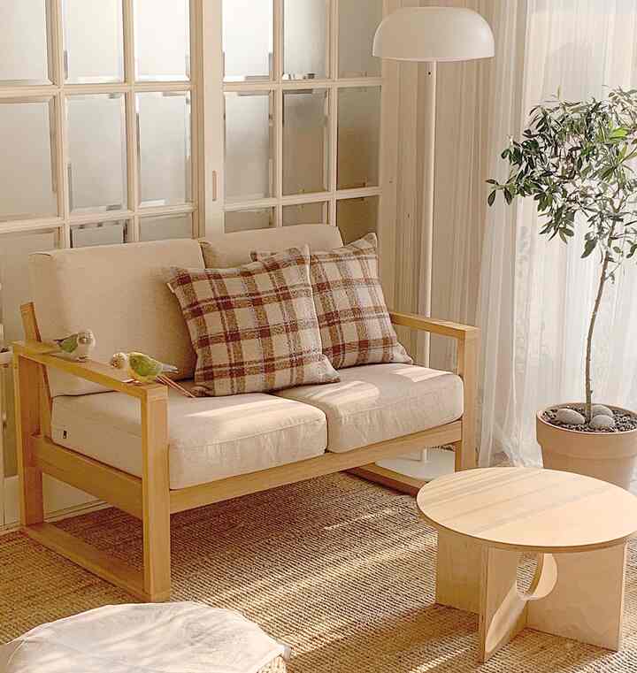 A living room featuring wood tones and white curtains, with a wooden sofa, woven rug, and a plant creating a natural atmosphere.