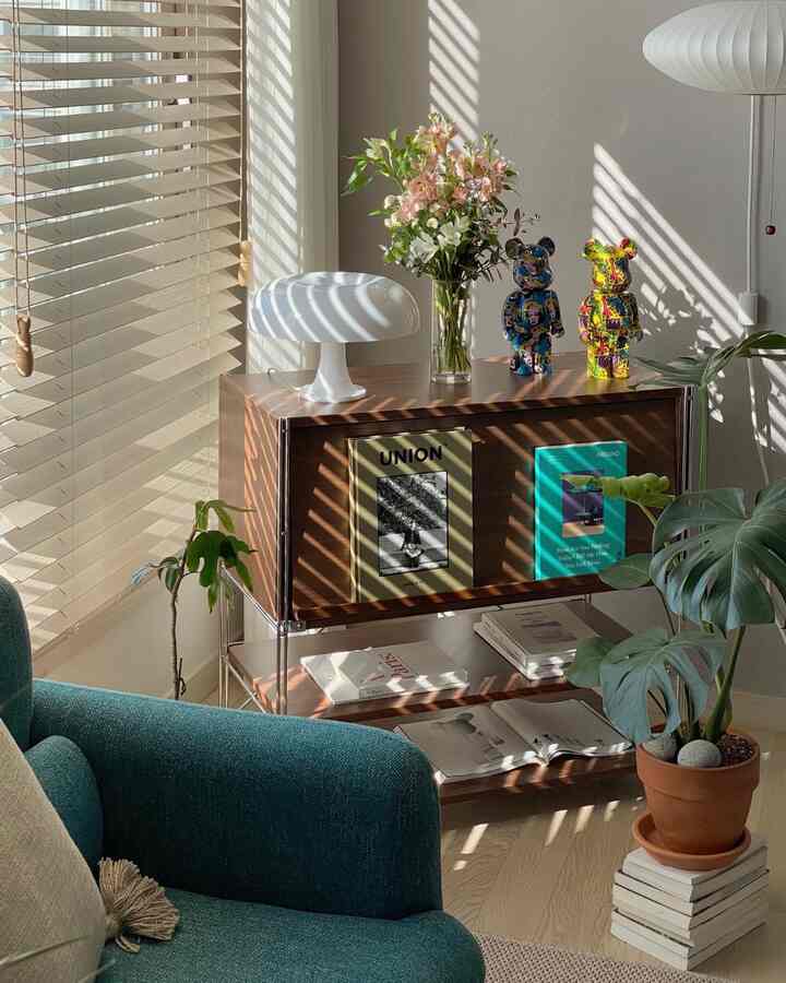 A warm and cozy living room featuring wood tones, natural sunlight filtering through blinds, plants, and a table lamp